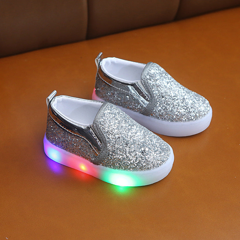 Silver sequin kids LED sneakers with light-up soles on brown surface.