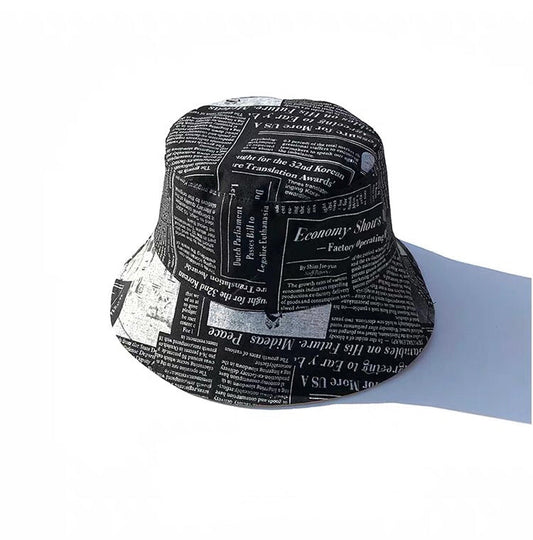 Reversible Newspaper Print Bucket Hat with white newspaper-style lettering on a black background, shown from an angled view.